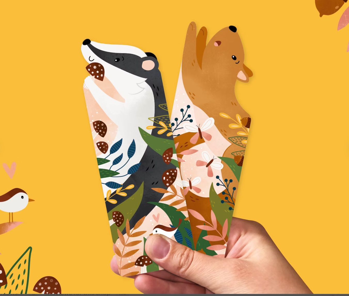 Squirrel and badger bookmarks being held by a white hand on a yellow background. 