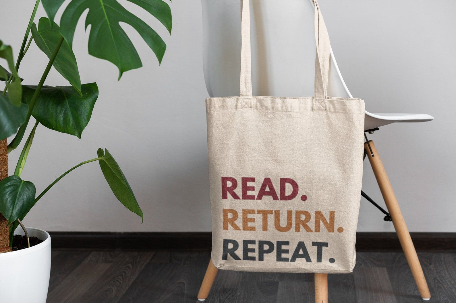 Image of a canvas tote bag with the words "read. return. repeat." The bag is on the back of a white chair and beside a big plant. 