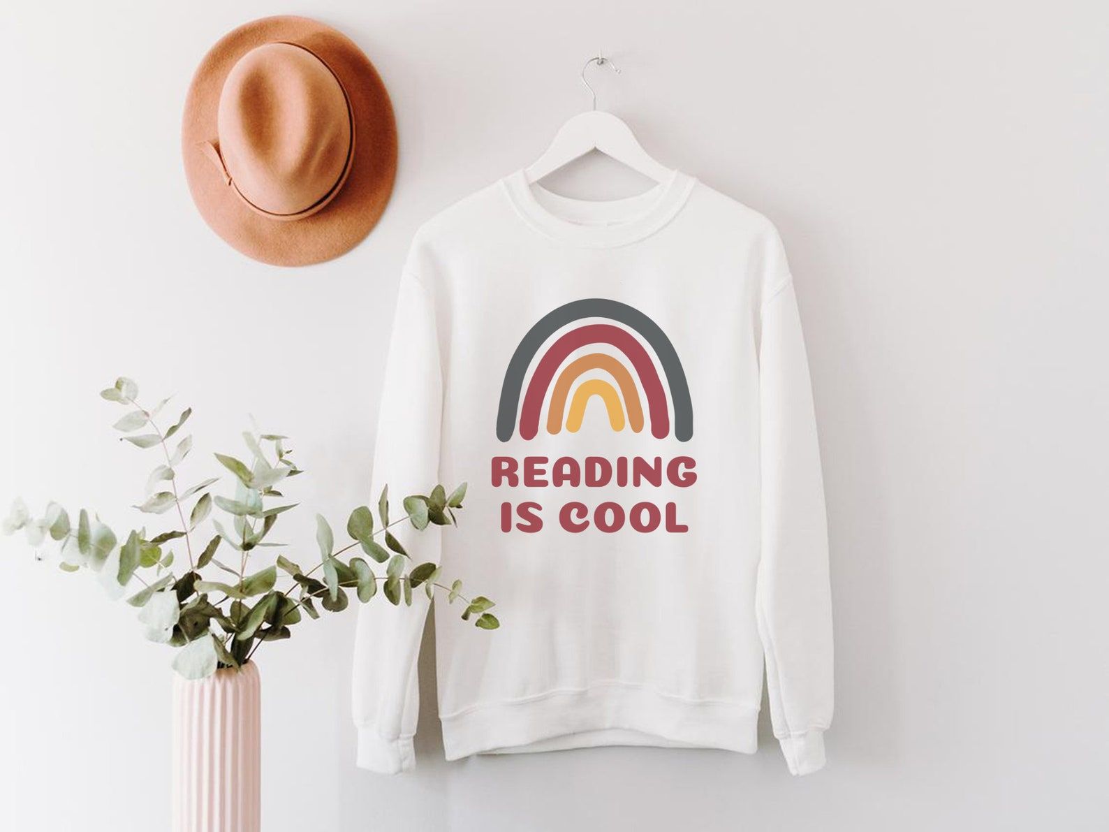 Image of a white sweatshirt with a rainbow and the words "reading is cool." There is a plant in front of it. 