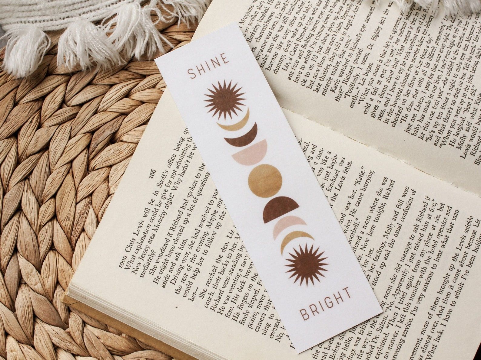 Image of a white bookmark on an open book. The bookmark reads "Shine Bright," and between the words are the moon phases. 