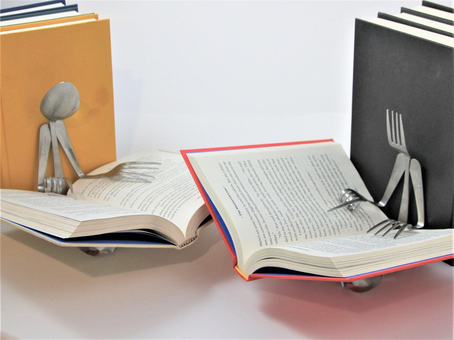 Set of bookends. The one on the left is a spoon, using forms to hold open a book. The right is a fork, also using forks to hold open a book. 