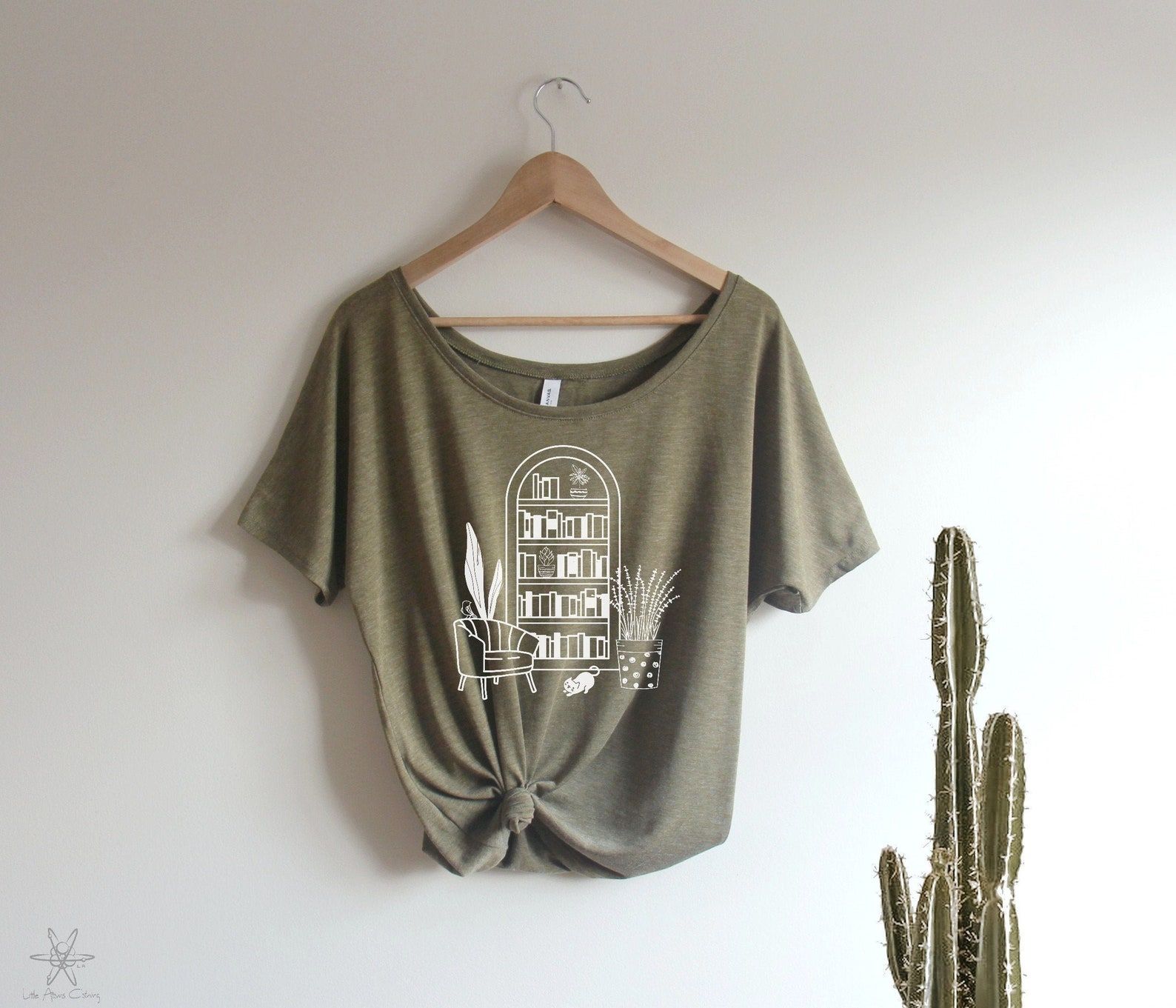 Sage green shirt with a rounded bookshelf, a comfy chair, and plants. 