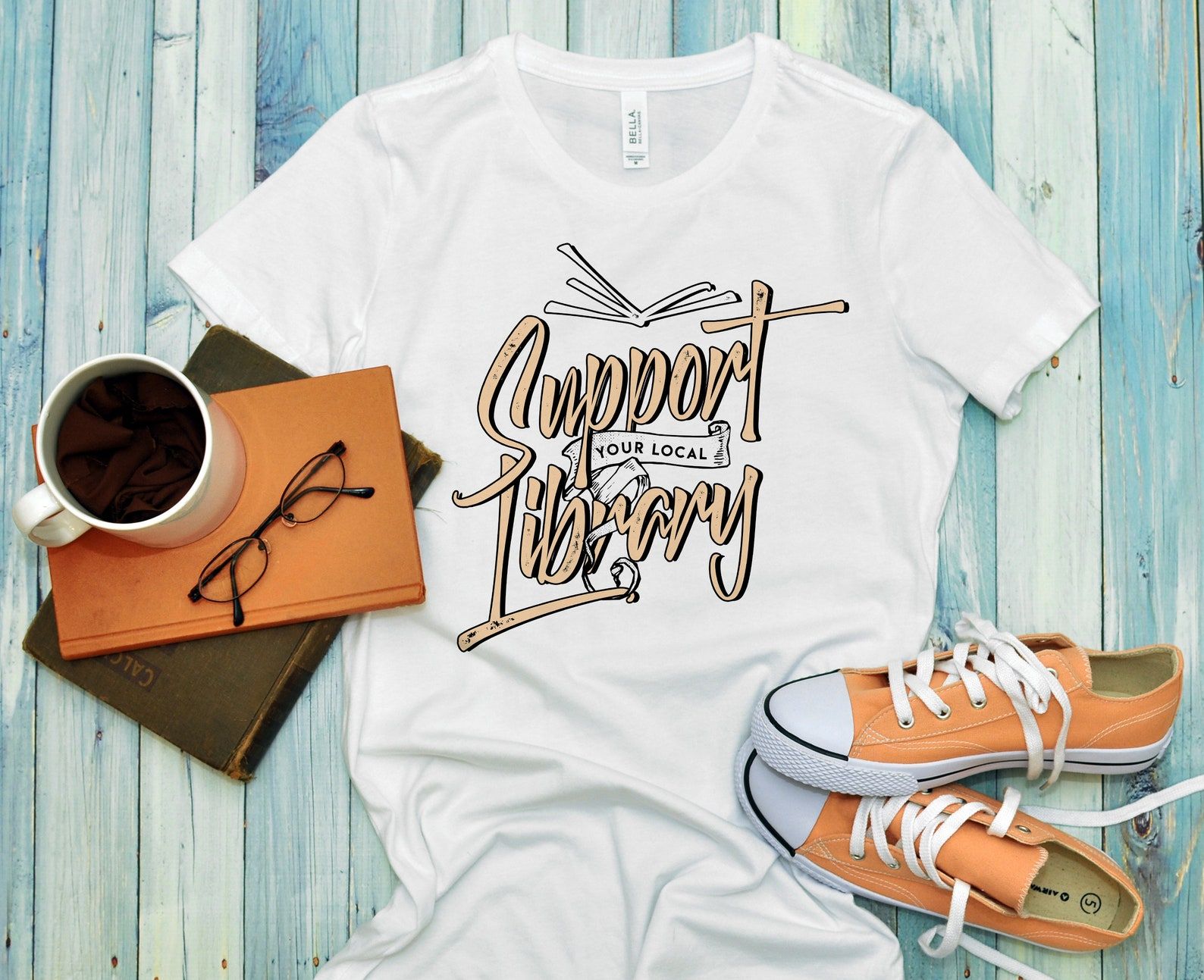Image of a white t-shirt which reads "support your local library." It's beside a stack of books and orange shoes. 
