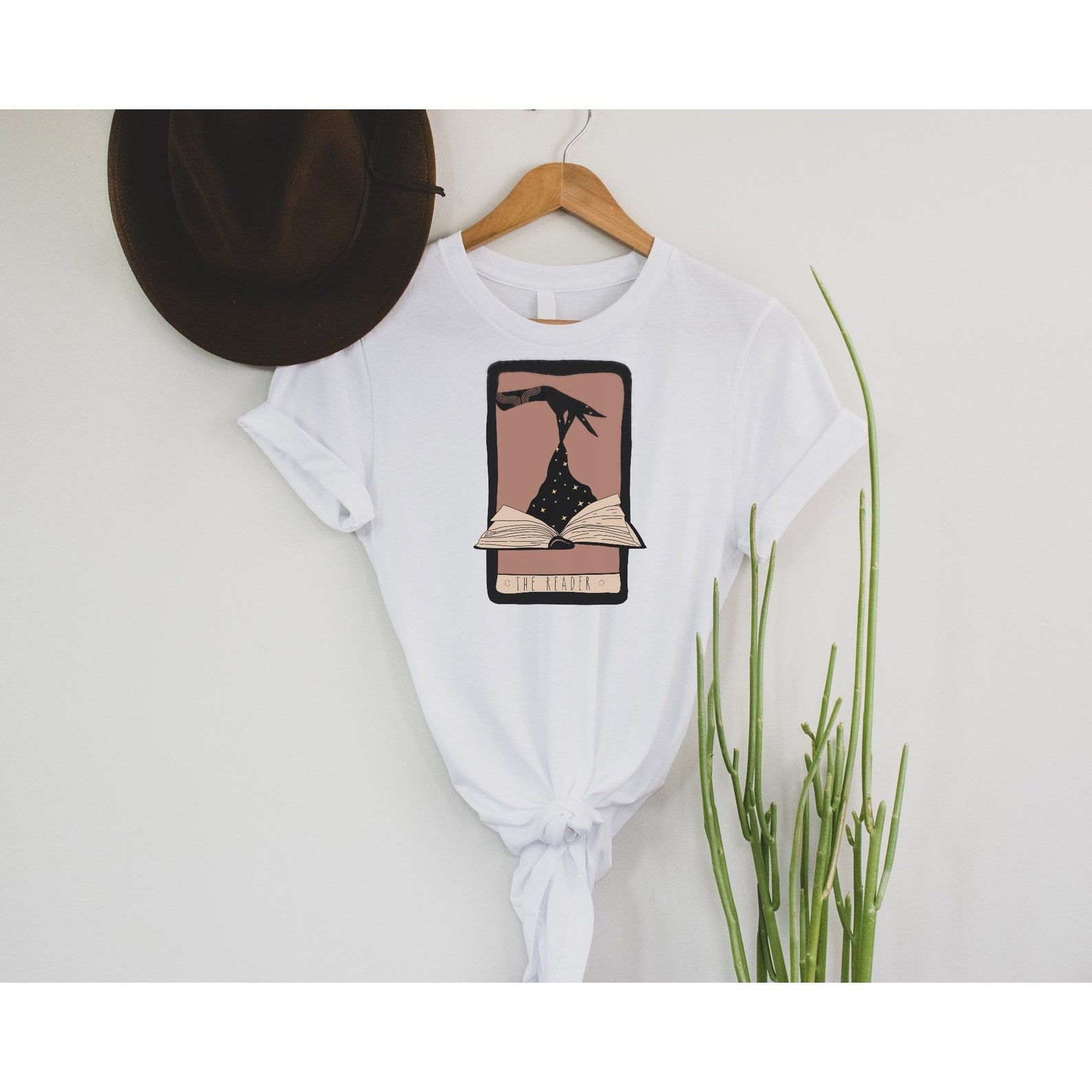 White t-shirt with a tarot card. On it is a white hand, pulling stars from an open book. The card is called "The Reader." 