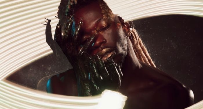 a Black male model with gold lipstick leaning his head into pair of shiny black hands with spikes for nails