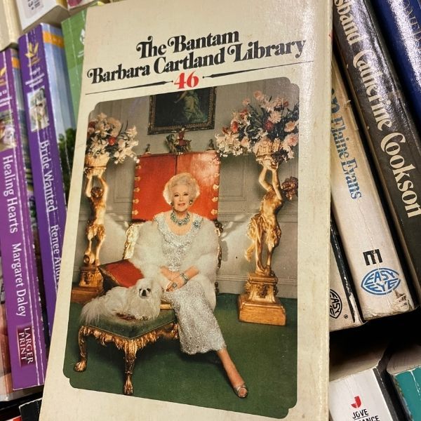 Le dos d'un roman de Barbara Cartland, avec des statues opulentes, un chien sur un pouf et l'auteur resplendissant de robe, de fourrure et de bijoux.