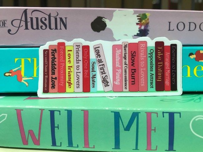 A 3" long by 1" tall sticker showing 16 colorful book spines that describe romance tropes. 