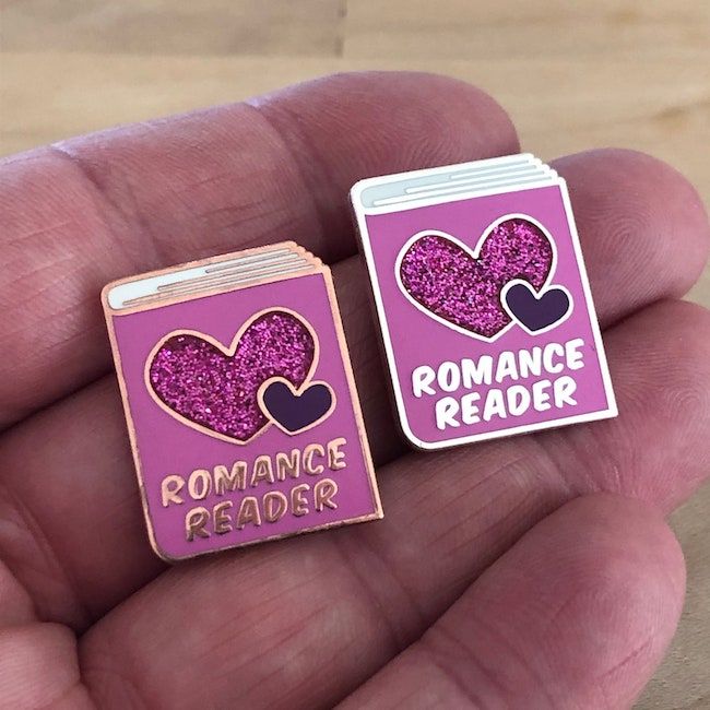 Two book-shaped enamel pins, pink with silver or rose gold highlights. The title of the book is "ROMANCE READER."