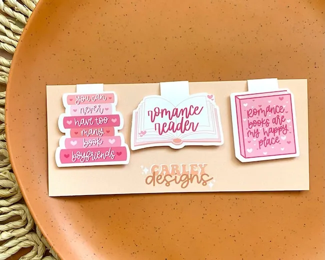 Three pink and white magnetic bookmarks shaped like books. The one on the left says, "you can never have too many book boyfriends." The middle says "romance reader" and one on the right says, "romance books are my happy place." 