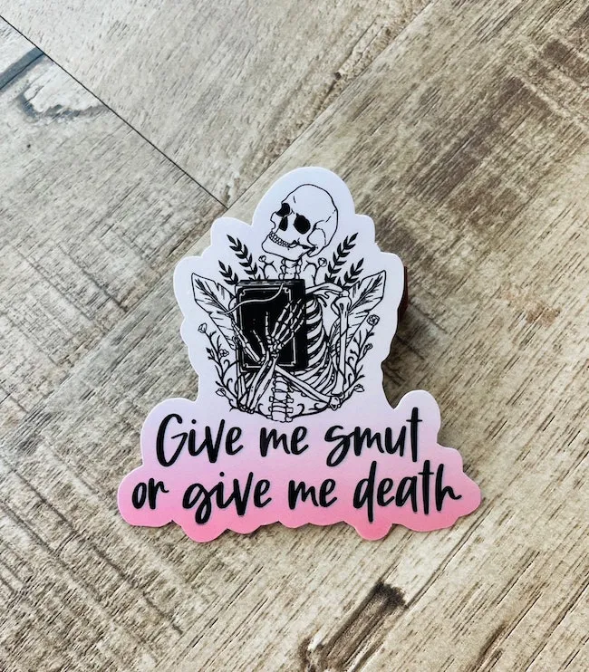 A gradient white-to-pink vinyl sticker with a skeleton holding a book and text that says "give me smut or give me death." 