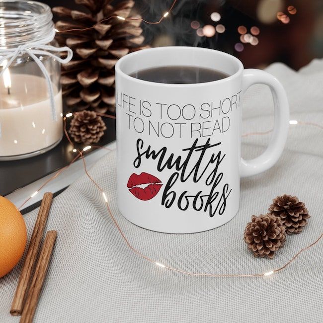 A white coffee mug that reads "life is too short to not read smutty books" in black lettering with a red lipstick kiss. 