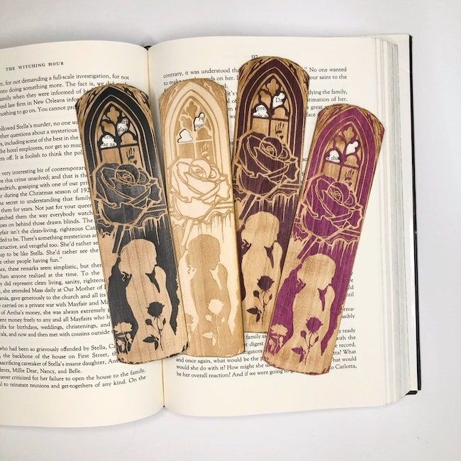 Four etched wooden bookmarks in black, natural, dark red, and fuchsia; all have the same design. It is the silhouette of a woman surrounded by roses under a Gothic window. 