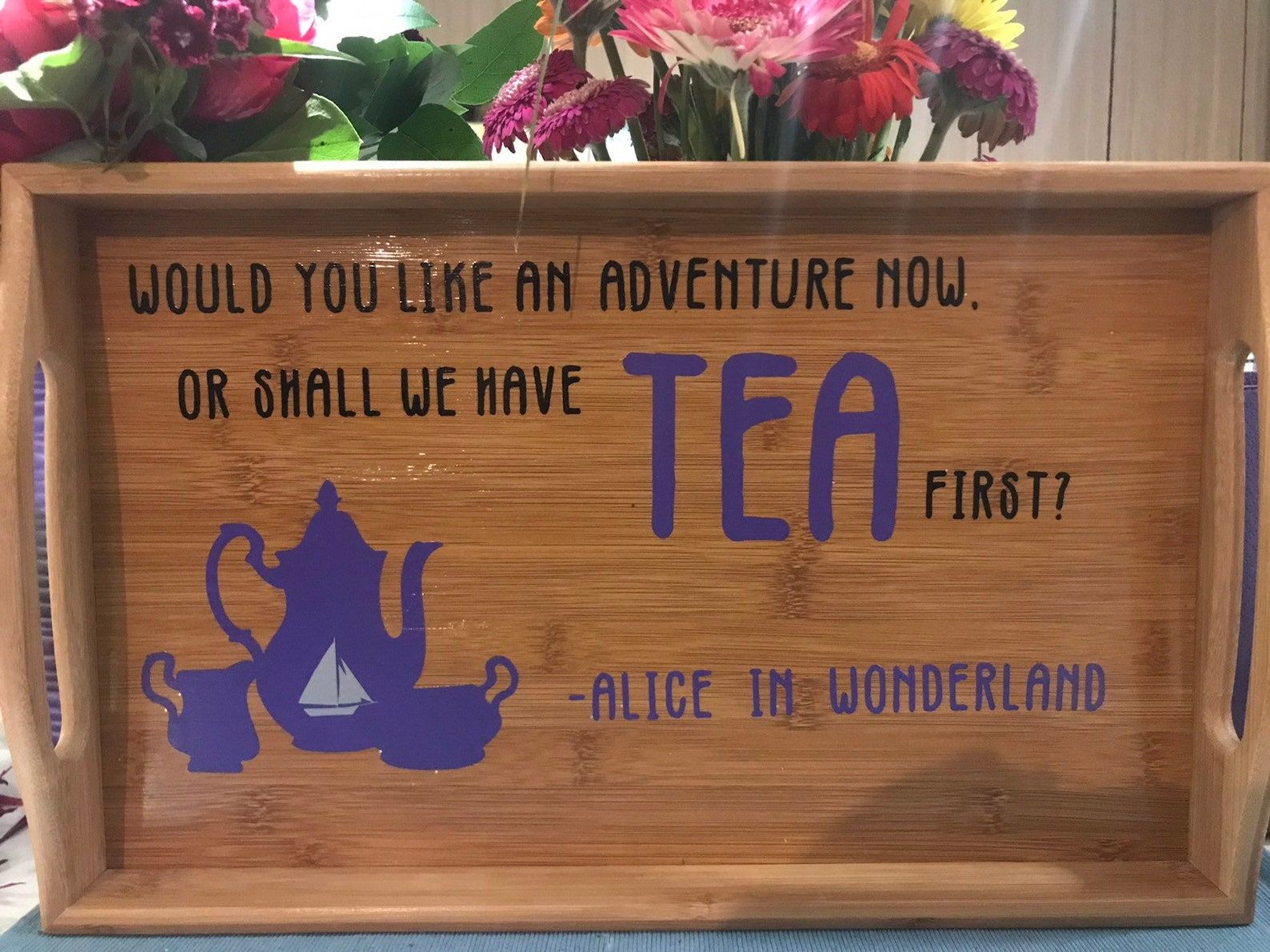 An overhead shot of a light brown wood serving tray. The words, "Would you like an adventure now, or shall we have tea first? - Alice in Wonderland." In the bottom left hand corner of the serving tray is a graphic of a tea spot, milk jug, and sugar container. 