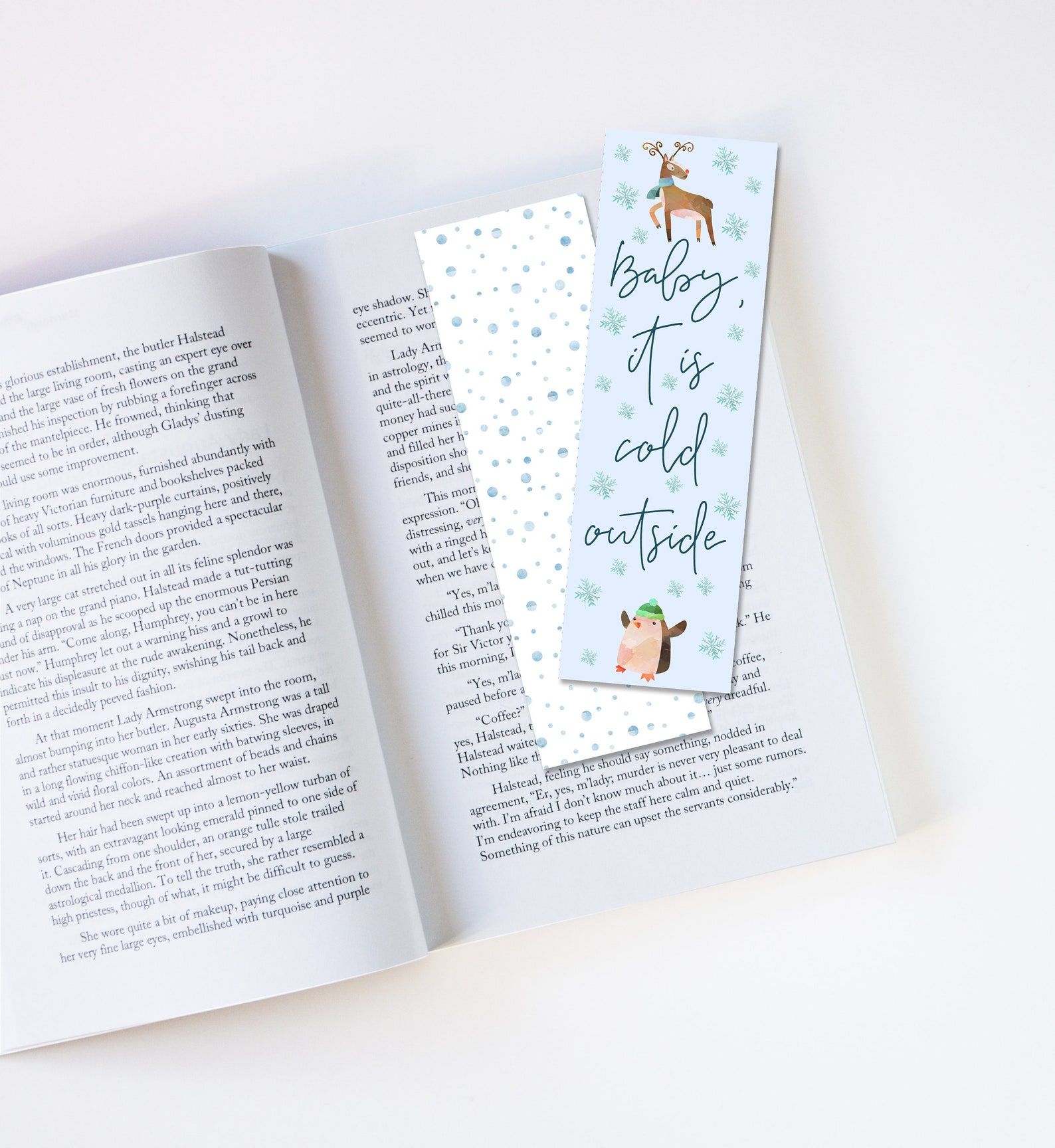 Image of the front and back of a bookmark on top of an open book. The back of the bookmark is white with blue snow. The front of the bookmark reads "Baby it's cold outside" in script, with a deer and penguin image. 