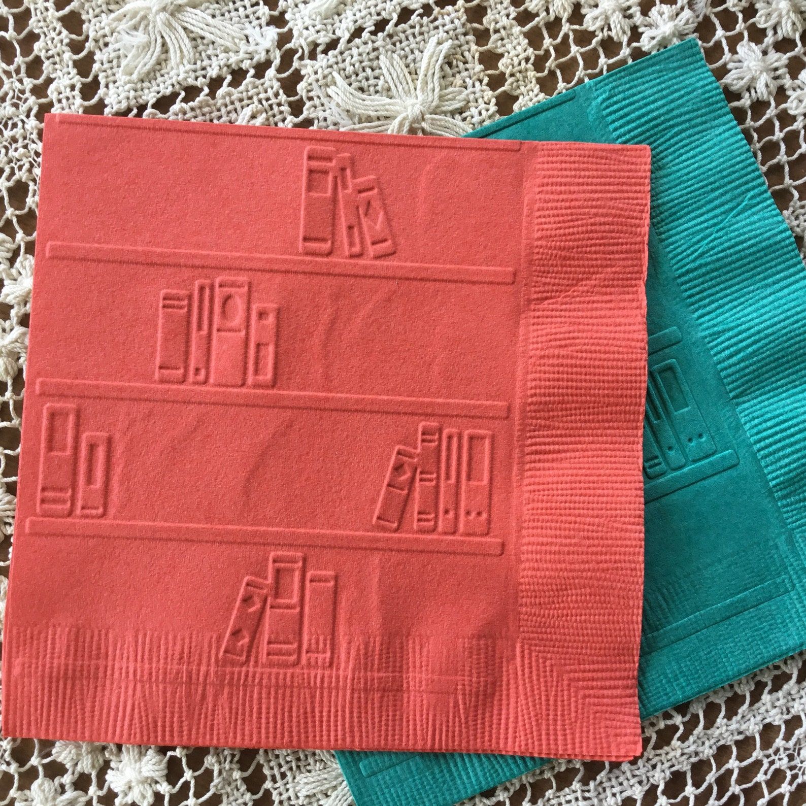 A red napkin and a teal napkin lie on top of a white doily. The napkins are the disposable paper ones with a graphic of a bookshelf embossed in. There are four levels of the bookshelf with different numbers of books on the respective shelves.