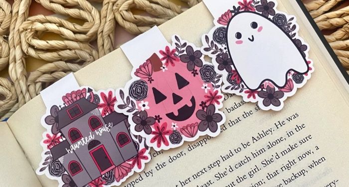 An open book with three magnetic bookmarks: a grey haunted house with pink florals, a pink jack-o-lantern, and a white ghost with pink florals.