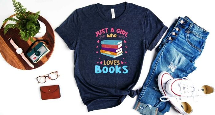 A flatlay centered around a navy t-shirt that reads "Just a girl who loves books" with colorful book spines and hearts, surrounded by jeans, sneakers, glasses, a watch and camera and a wallet.