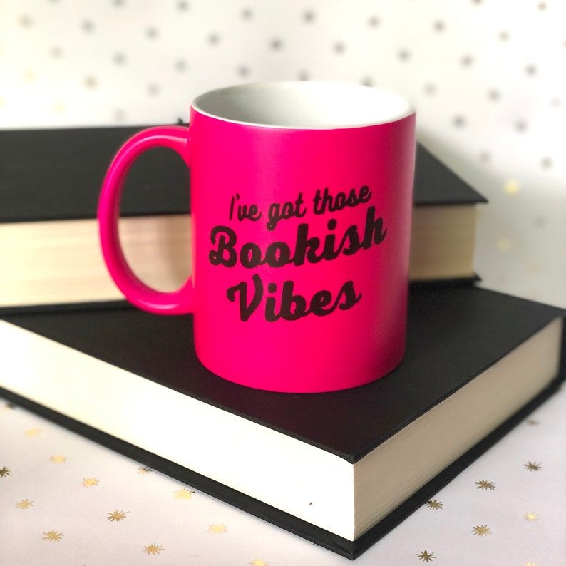 Bright pink mug with the words "I've got those bookish vibes" in a retro black font. It's sitting on top of a black hardcover book. 