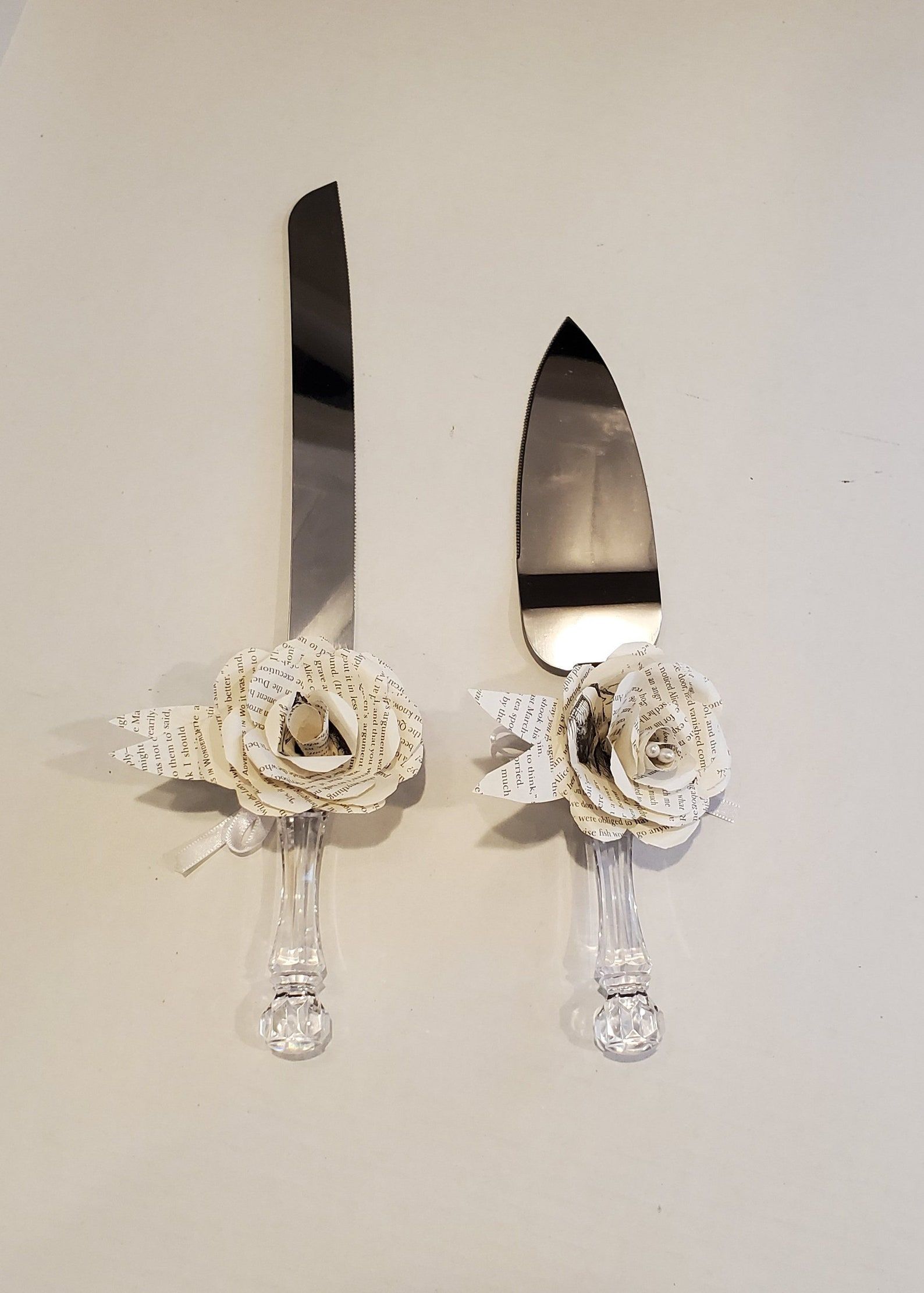 An overhead shot of a cake knife and a cake server. They are plated server with clear handles. On the necks of both utensils is a paper flower made out of pages of books 