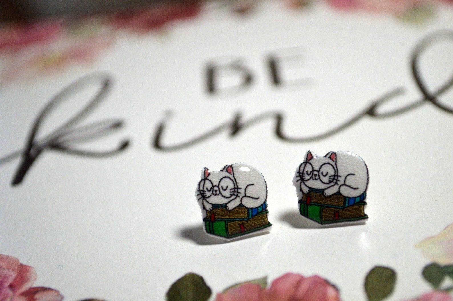 Earrings of tiny white cats wearing glasses sleep on a stack of books.