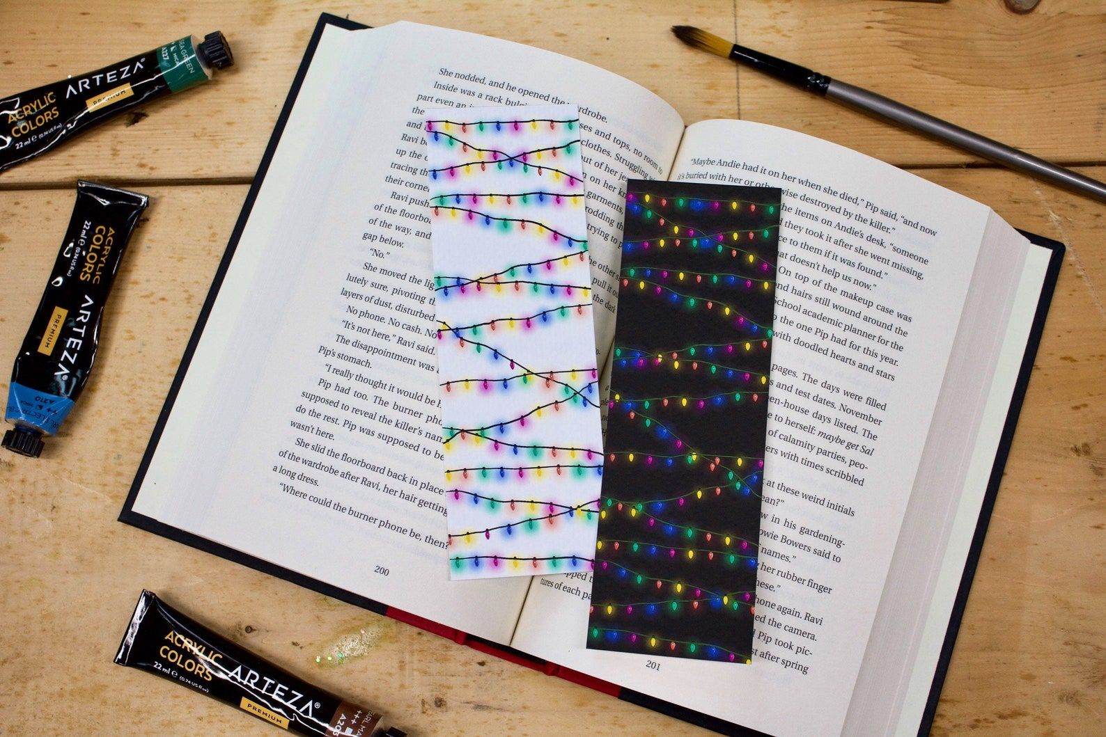 Image of two bookmarks featuring Christmas lights. One is black with colorful lights and one is white with colorful lights. They are sitting on top of an open book. 