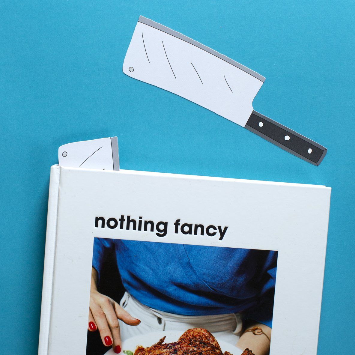 A bookmark in the shape of a cleaver against a blue background, next to a cookbook