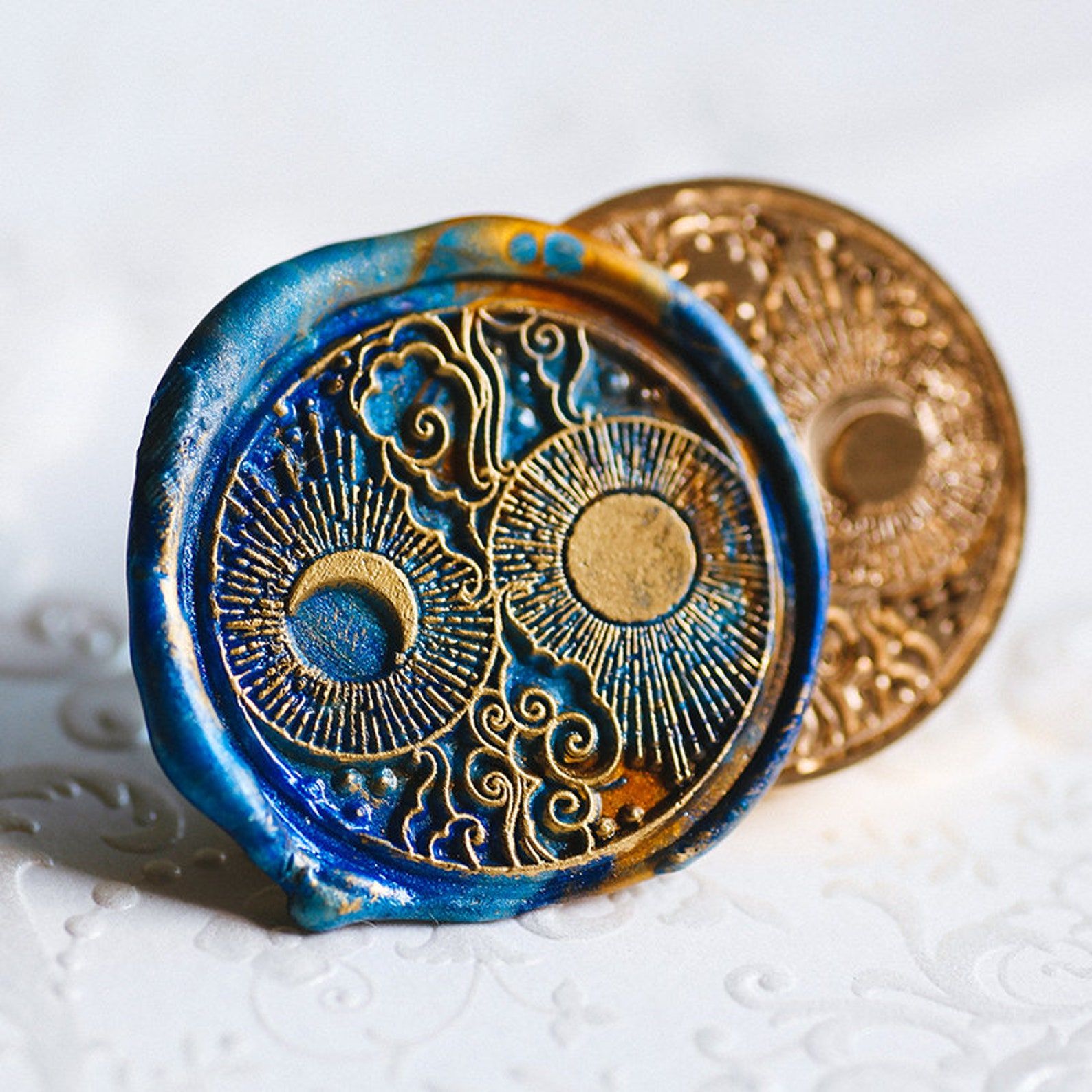 Image of a wax seal that is dark blue and gold. The seal has a sun, moon, and clouds. 