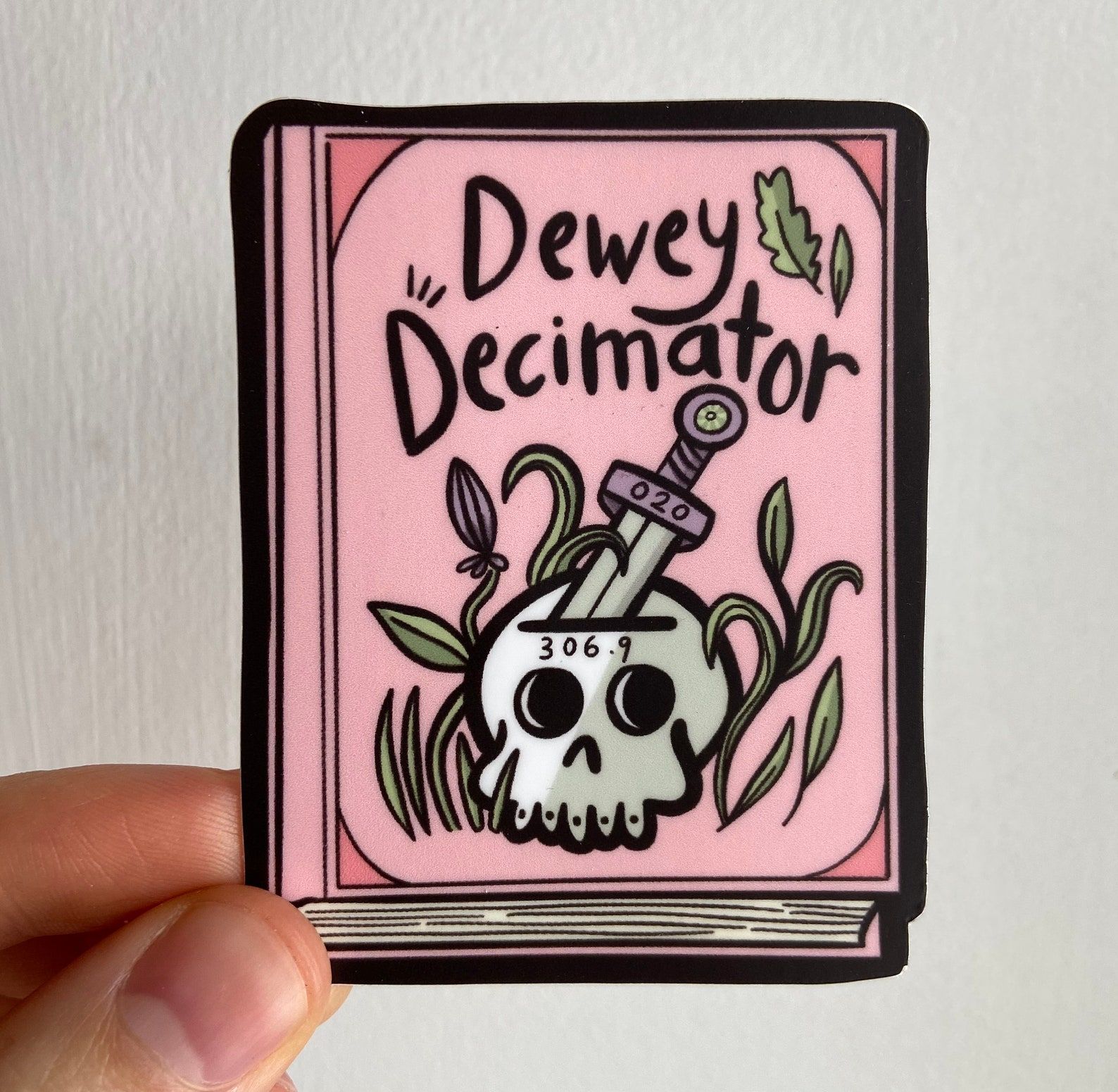 Image of a pink sticker in the shape of a book. On it is a skull and the words "dewey decimator." 