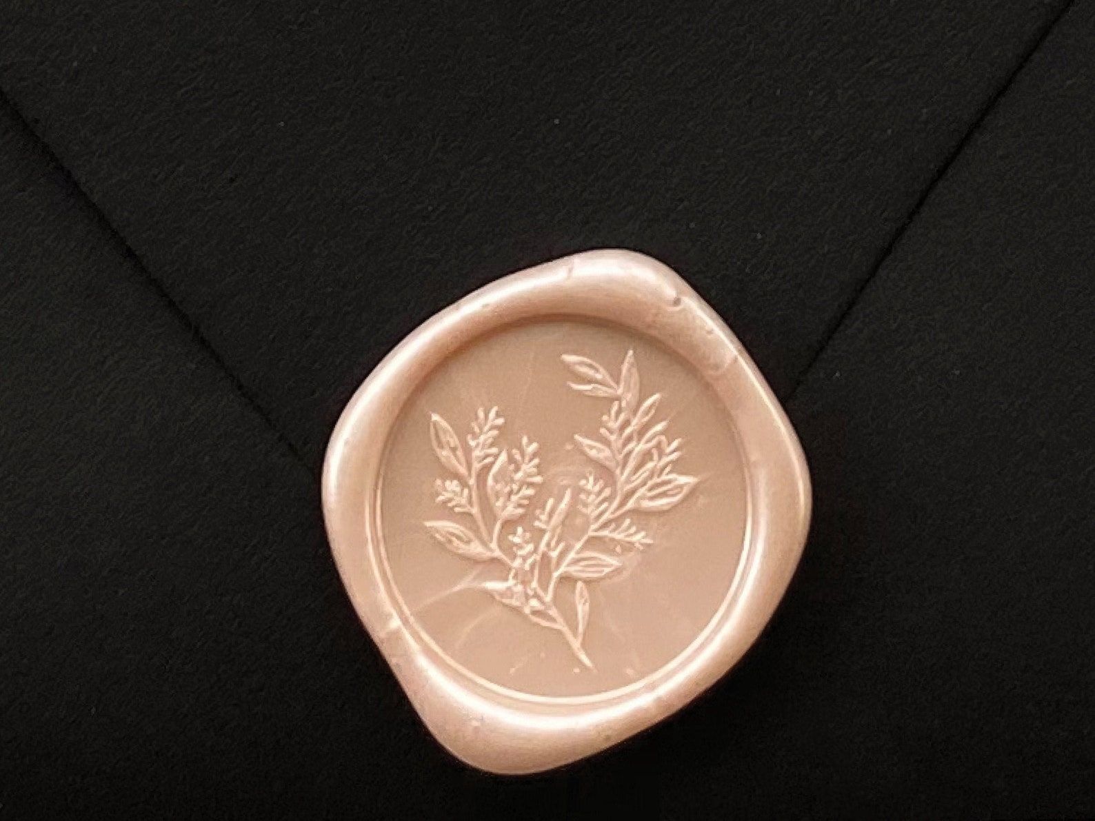 Image of a peach colored wax stamp on a black envelope. The stamp has a eucalyptus branch in the center. 