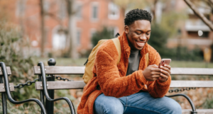 A young Black man sitting on a bench, wearing an orange fleece jacket and smiling down at his phone.
