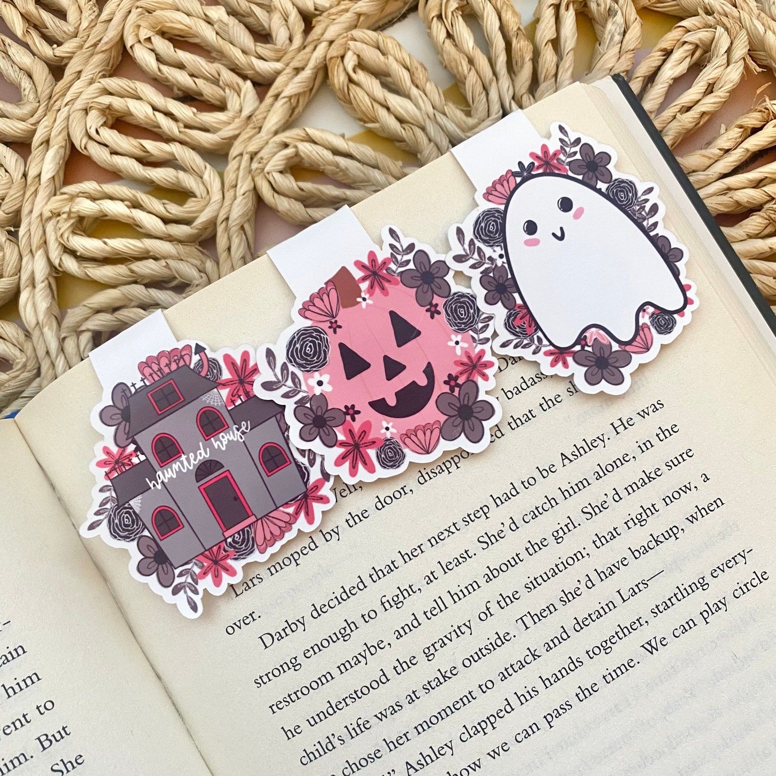 An open book with three magnetic bookmarks: a grey haunted house with pink florals, a pink jack-o-lantern, and a white ghost with pink florals.