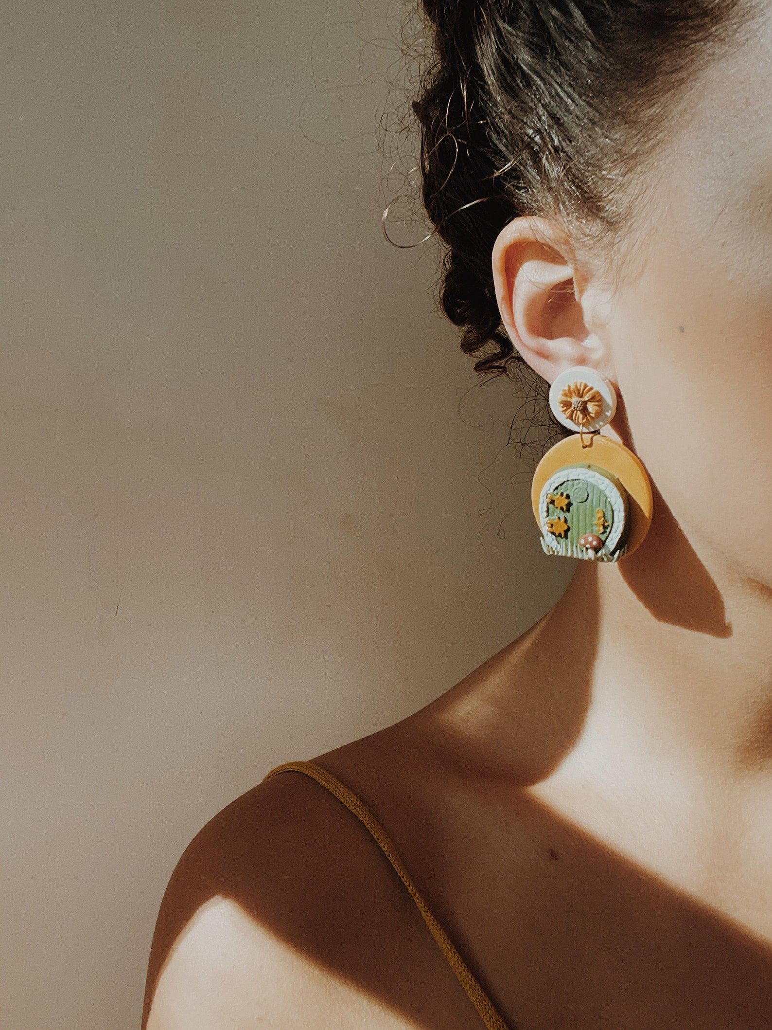 A photo of a woman wearing dangly earrings that look like a round green hobbit house door, with tiny hinges and mushrooms