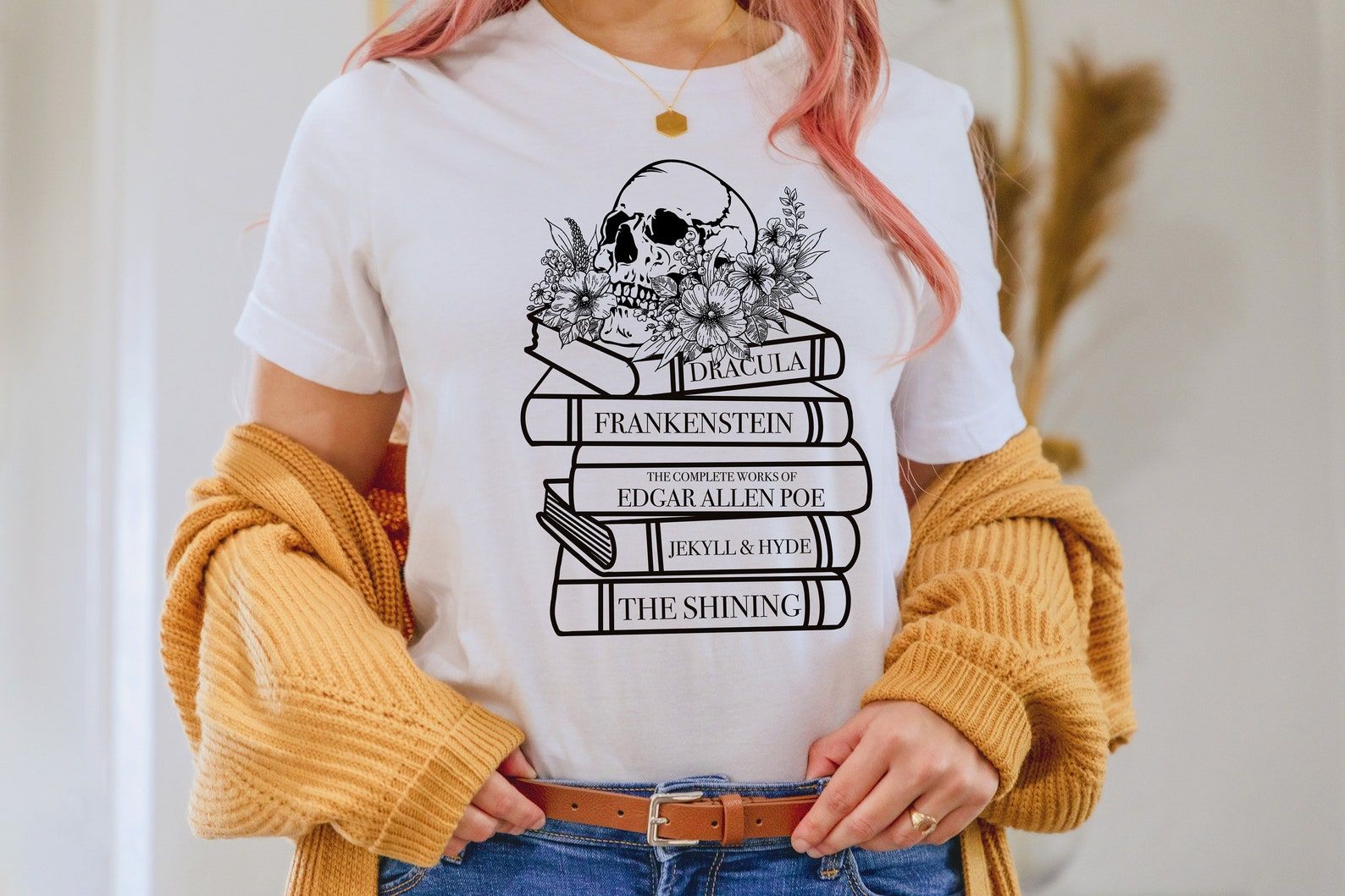 A white t-shirt with a stack of book spines featuring Dracula, Frankenstein, The Shining, Edgar Allan Poe, Jekyll and Hyde, with a skull and flowers on toy.