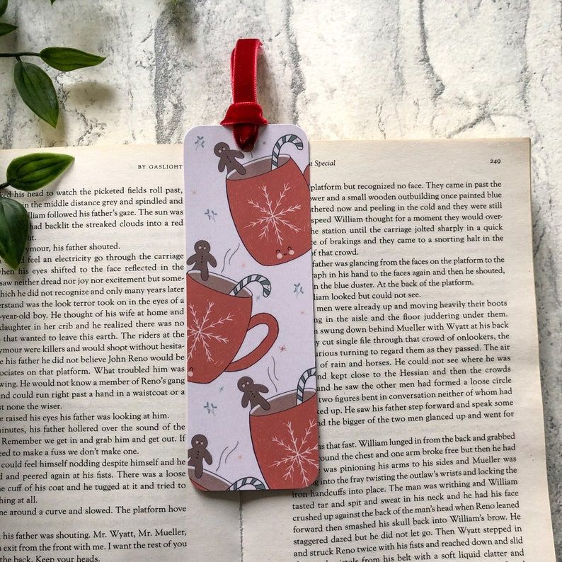 Image of a bookmark on an open book. The bookmark features coffee mugs with snowflakes, as well as gingerbread men and candy canes. 