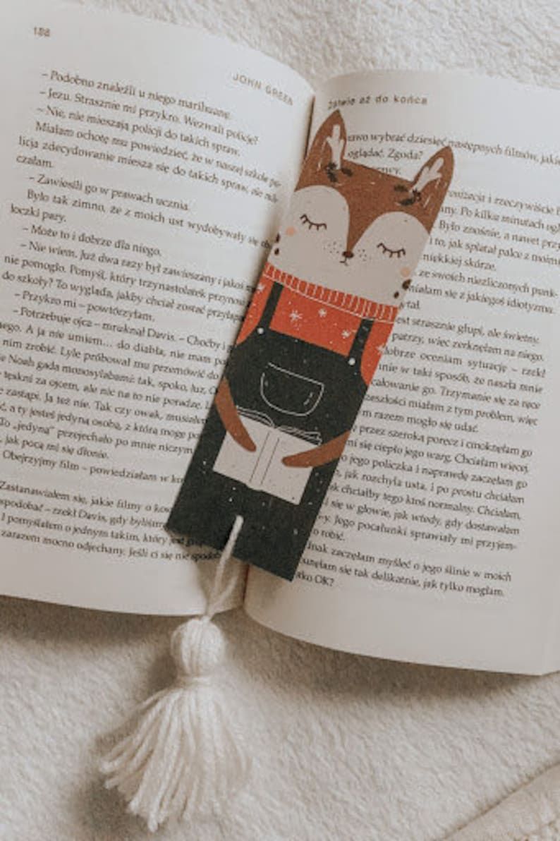 Image of a bookmark featuring a reindeer wearing overalls and reading a book. There is a white tassel on the bottom. 