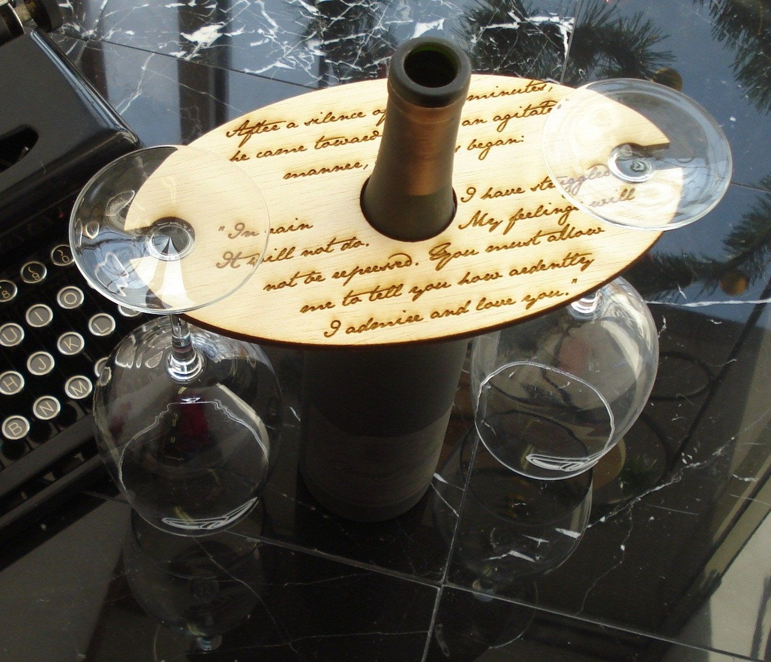 An overhead photo of a wine bottle and two wine glasses in a display. The two glasses can be stored upside down in this wine display and the board has a hole in the middle that can rest around the bottleneck of the wine bottle. There is a long quote from Pride and Prejudice engraved in.