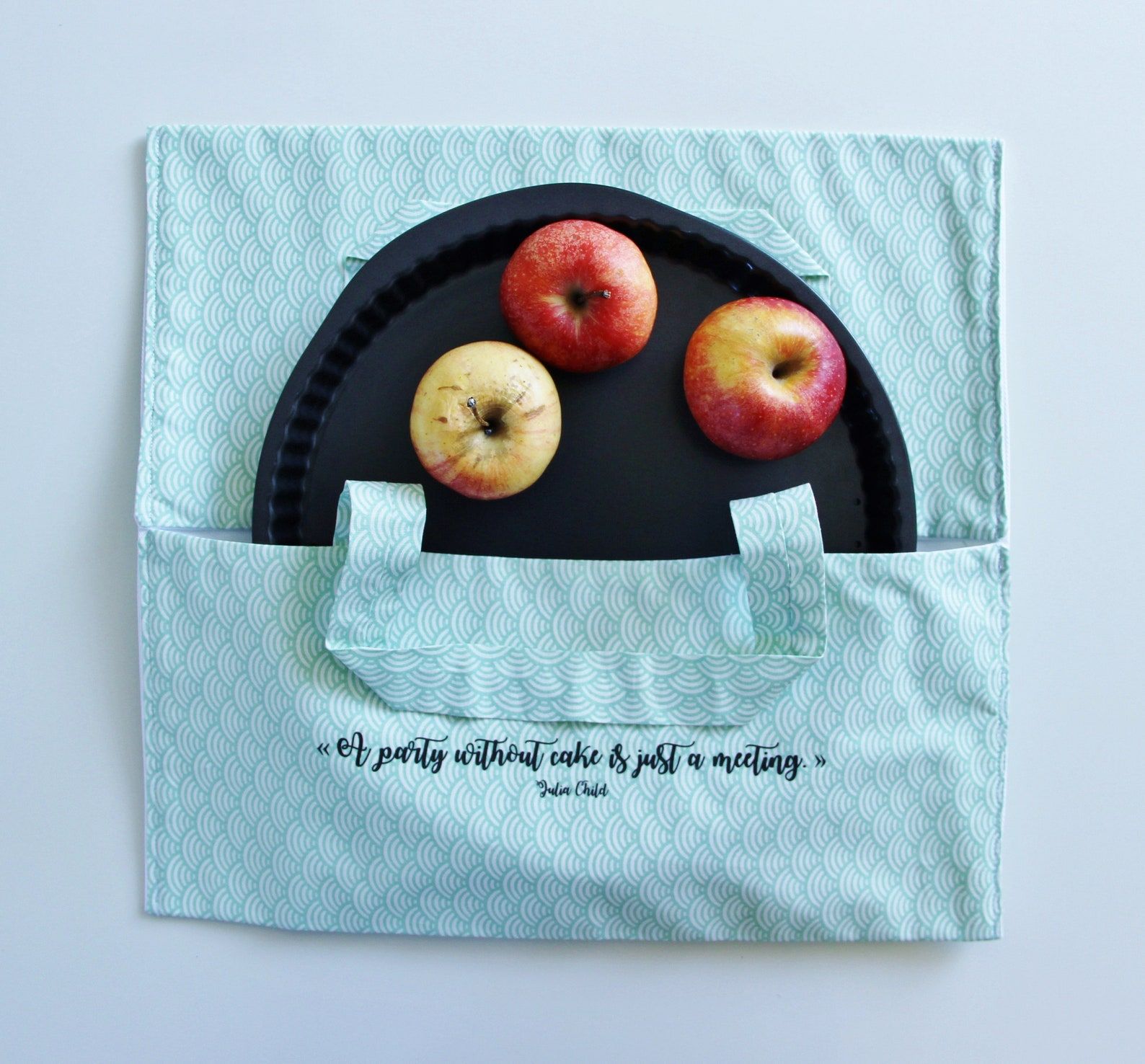 An overhead shot of a light blue, cloth pie carrier. There is a pie tin poking out of the carrier with three apples in it. There is writing on the bottom half of the pie carrier that reads, "A party without cake is just a meeting," Julia Child.