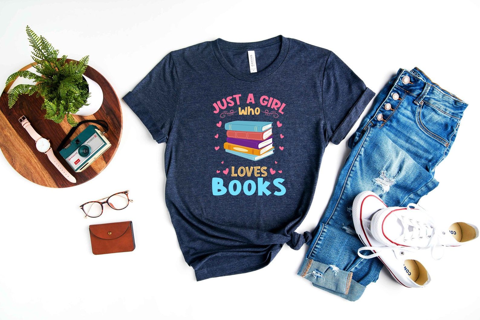 A flatlay centered around a navy t-shirt that reads "Just a girl who loves books" with colorful book spines and hearts, surrounded by jeans, sneakers, glasses, a watch and camera and a wallet.