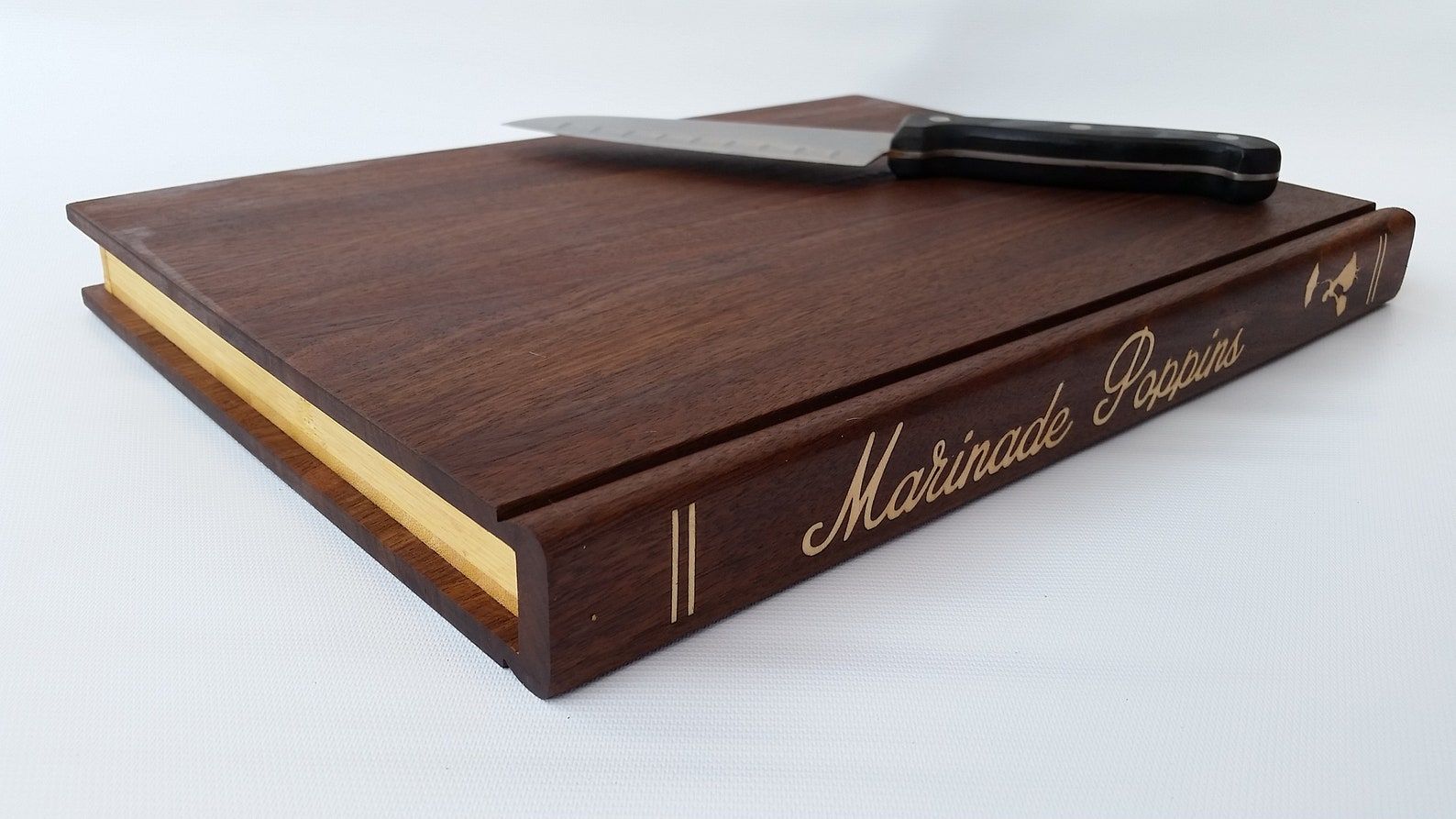 A deep brown cutting board in the shape of a book with a knife laying on the top right hand corner. The spine of the cutting board reads, "Marinade Poppins."