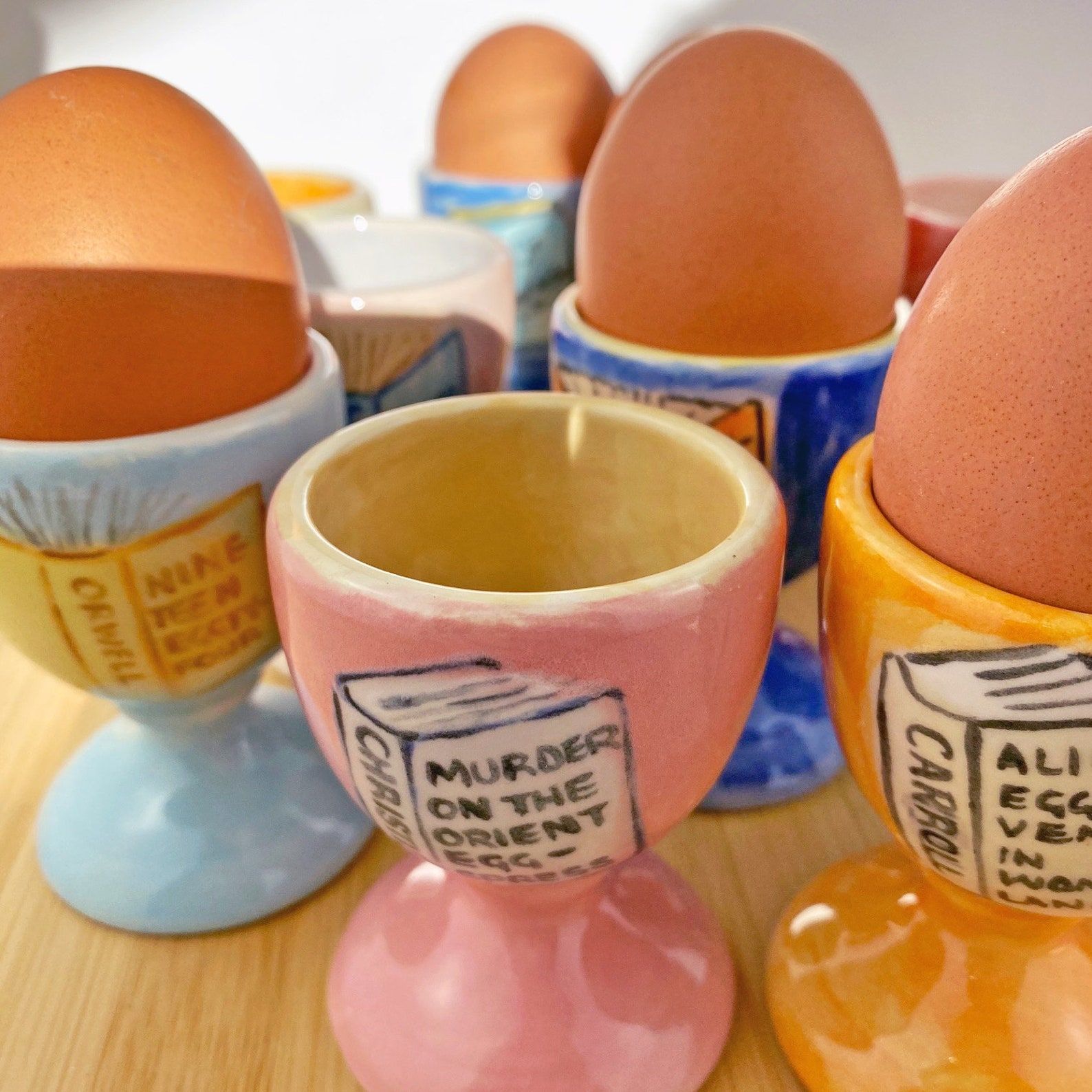 Une photo d'un groupe d'œufs peints avec des jeux de mots sur les titres de livres liés aux œufs.  Les coquetiers sont de différentes couleurs et peints à la main.