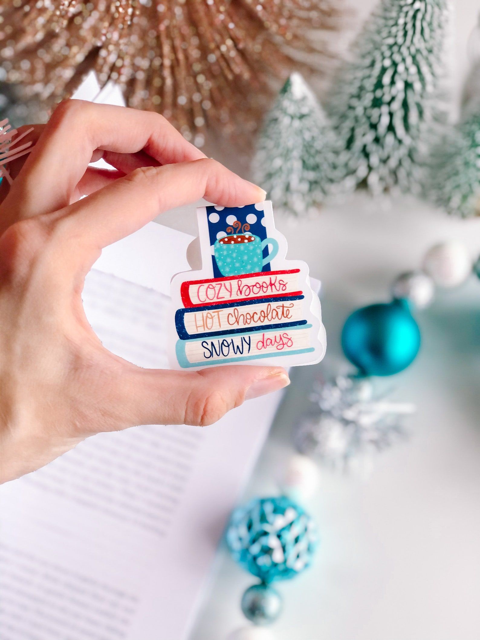 Image of a bookmark being held in front of blue and silver ornaments. The bookmark is magnetic and features a coffee mug on top of three books. The books read "cozy books, hot chocolate, snowy days." 