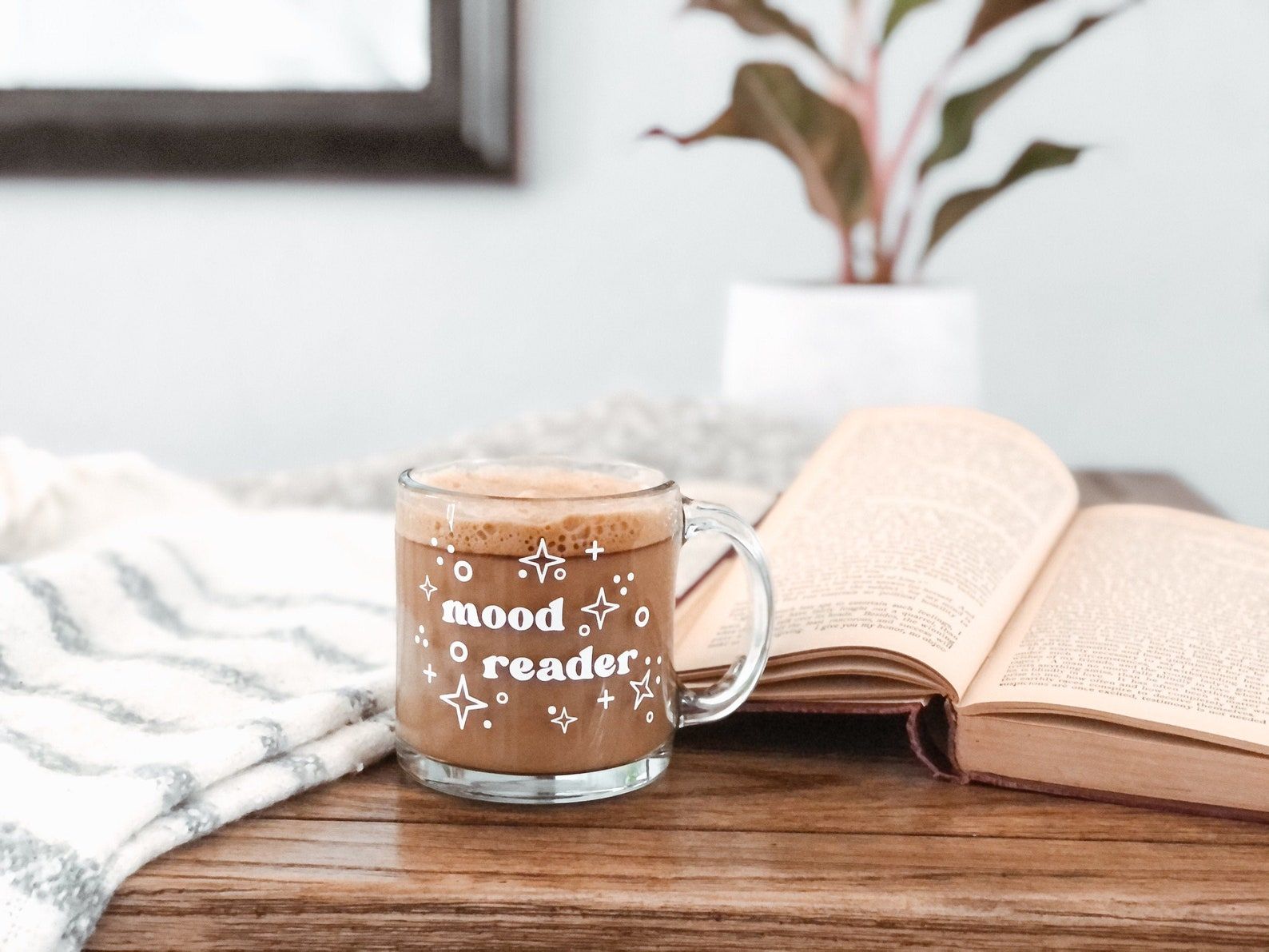 Glass mug with the words "mood reader" in white. The mug is sitting on a table beside a book and blanket. 