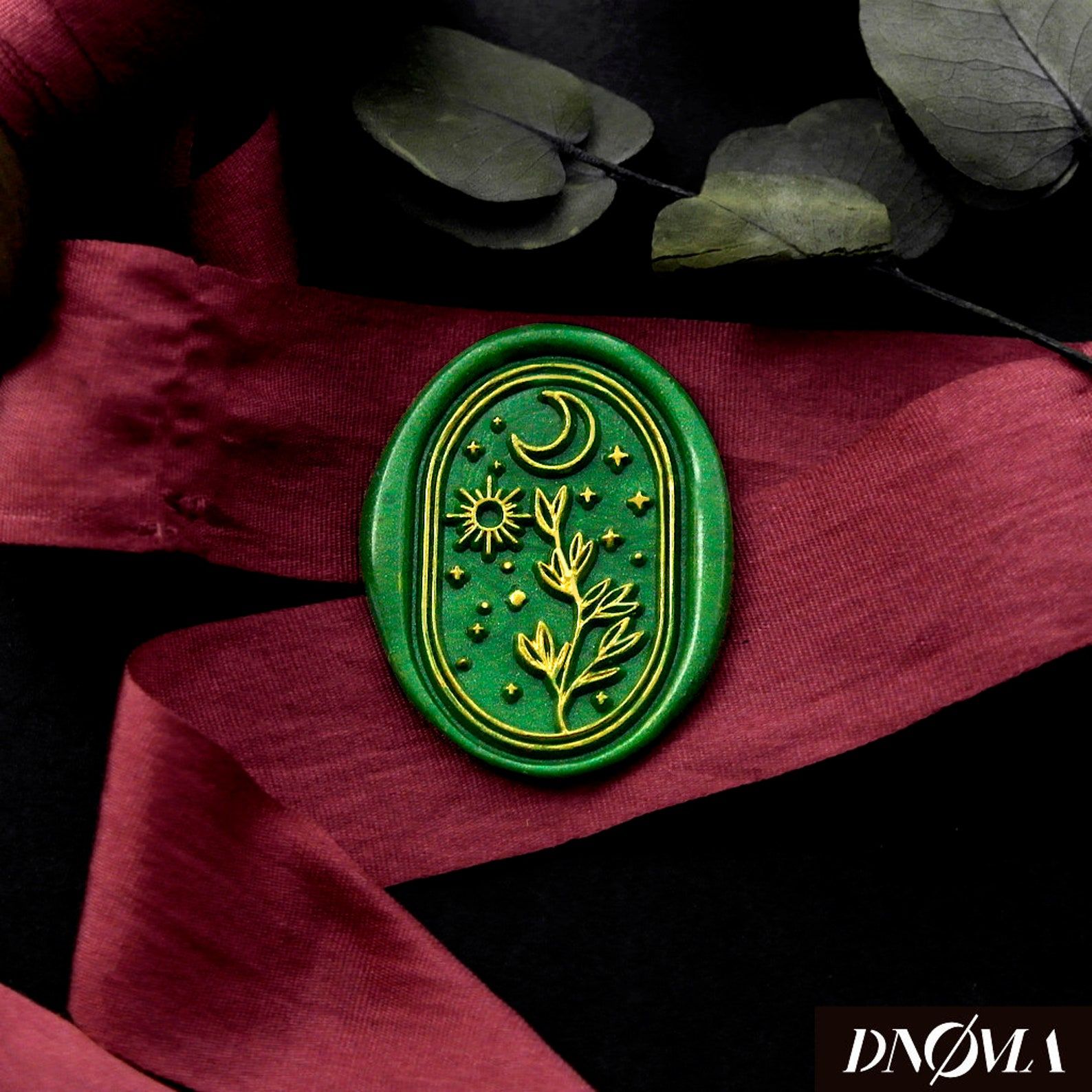 Image d'un sceau de cire vert foncé, représentant une lune, un soleil et une plante en or.  Ceux-ci sont en or et le timbre est au-dessus d'un ruban rouge.  