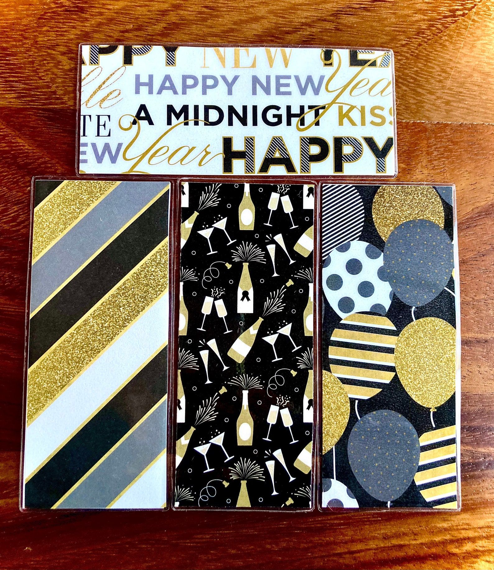 Image of three bookmarks on a table. The first bookmark is gold, silver, black, and white stripes. The second is black with gold and silver champaign bottles and glasses. The last bookmark features balloons in the same colorway. 