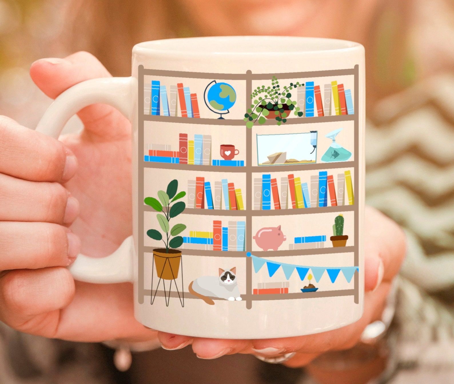 Two hands hold a peach-colored mug. The mug features colorful books on a set of shelves, with a cat, piggy bank, plant, and other decor. 
