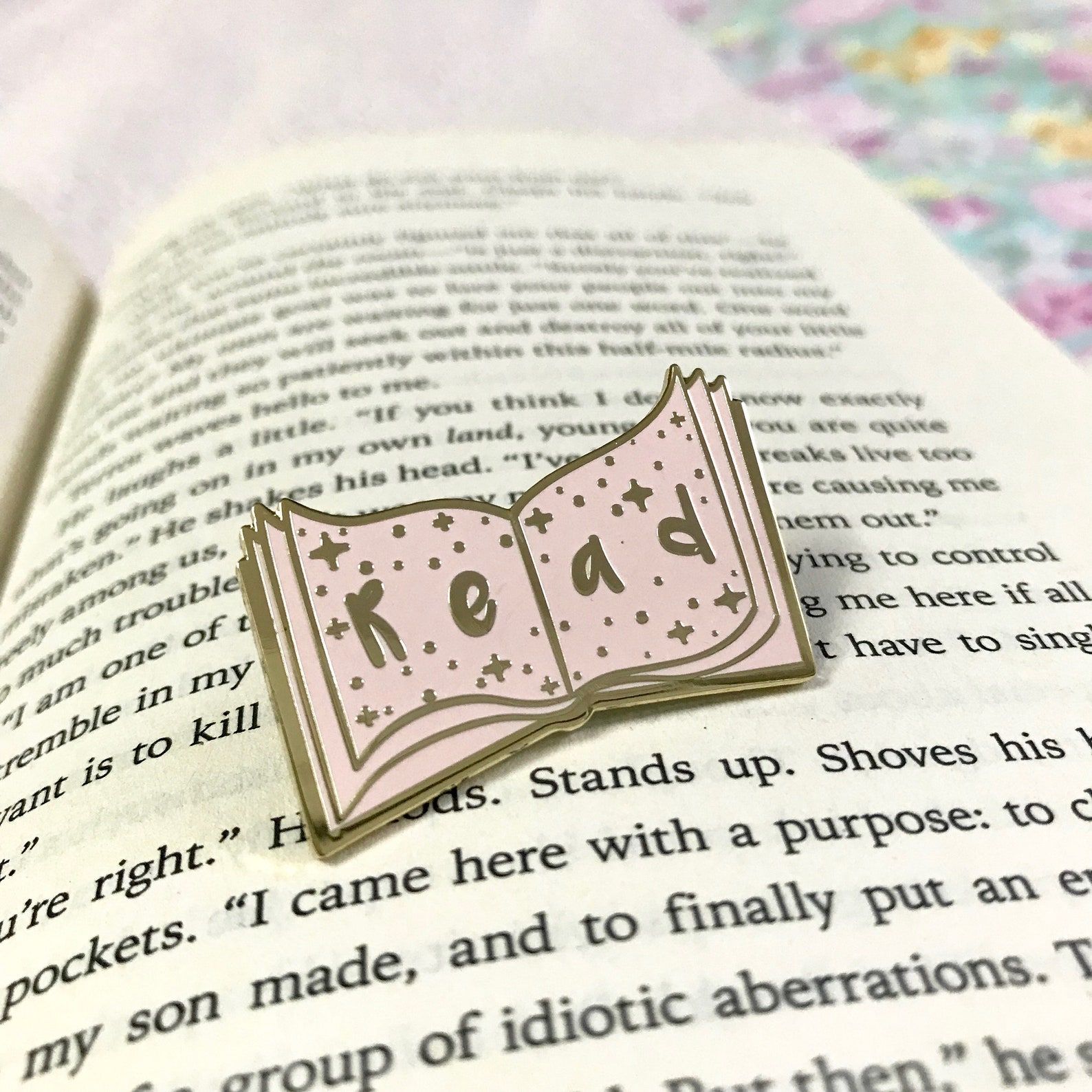 Pink enamel pin sitting on an open book. It reads 