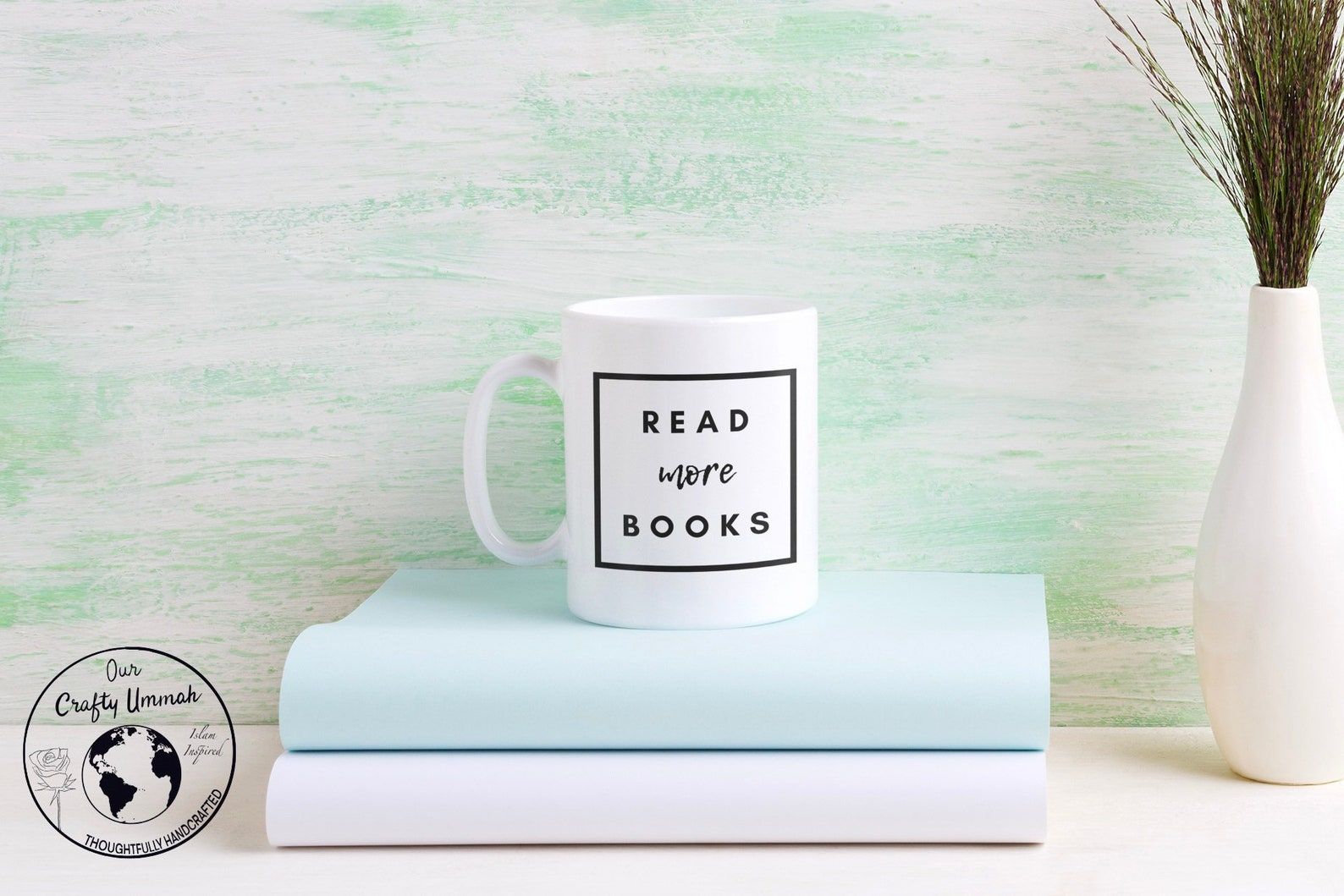 A white mug on a stack of books. The mug uses black font which reads "read more books." 
