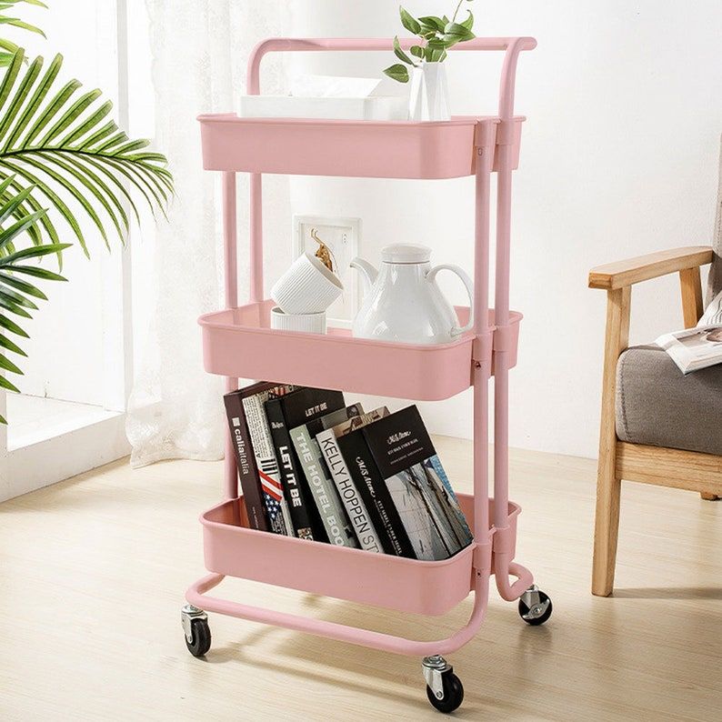 chariot à roulettes en métal rose avec des livres sur l'étagère inférieure et une théière sur l'étagère du milieu