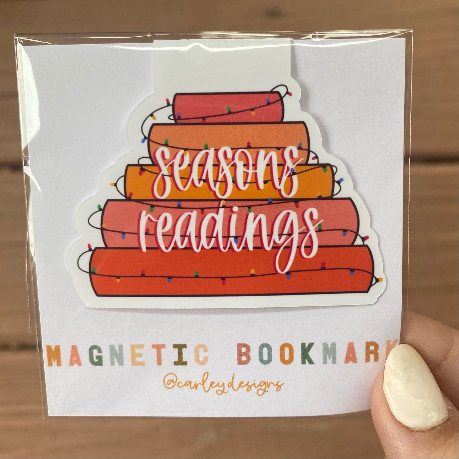 Image of a hand holding the package for a magnetic bookmark. The bookmark inside the package is a stack of books wrapped in lights and reads "Seasons readings" in a script font. 