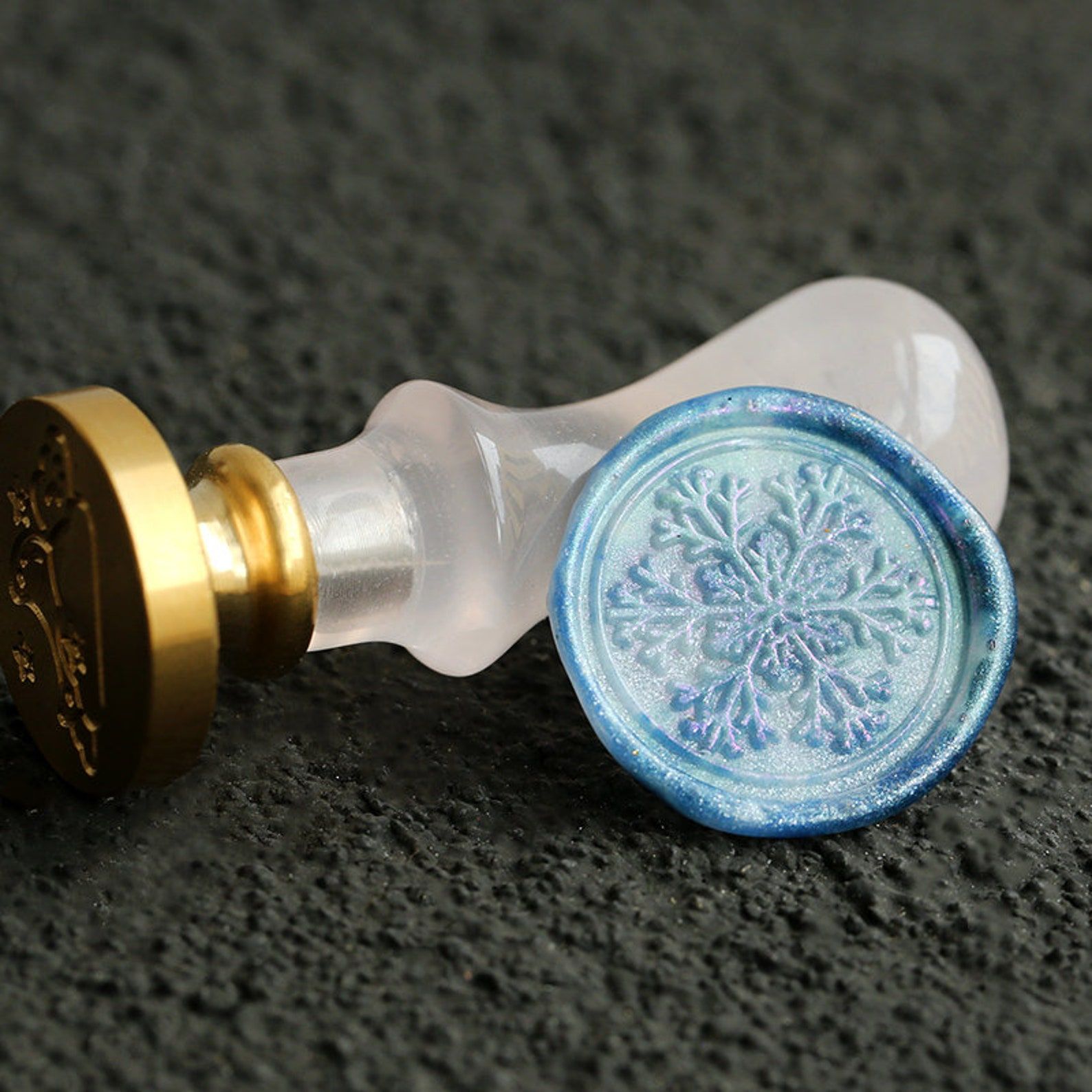 Image of a wax seal stamper with a white handle. The seal lies on top of it, a range of blue shades, with a snowflake in the middle. 